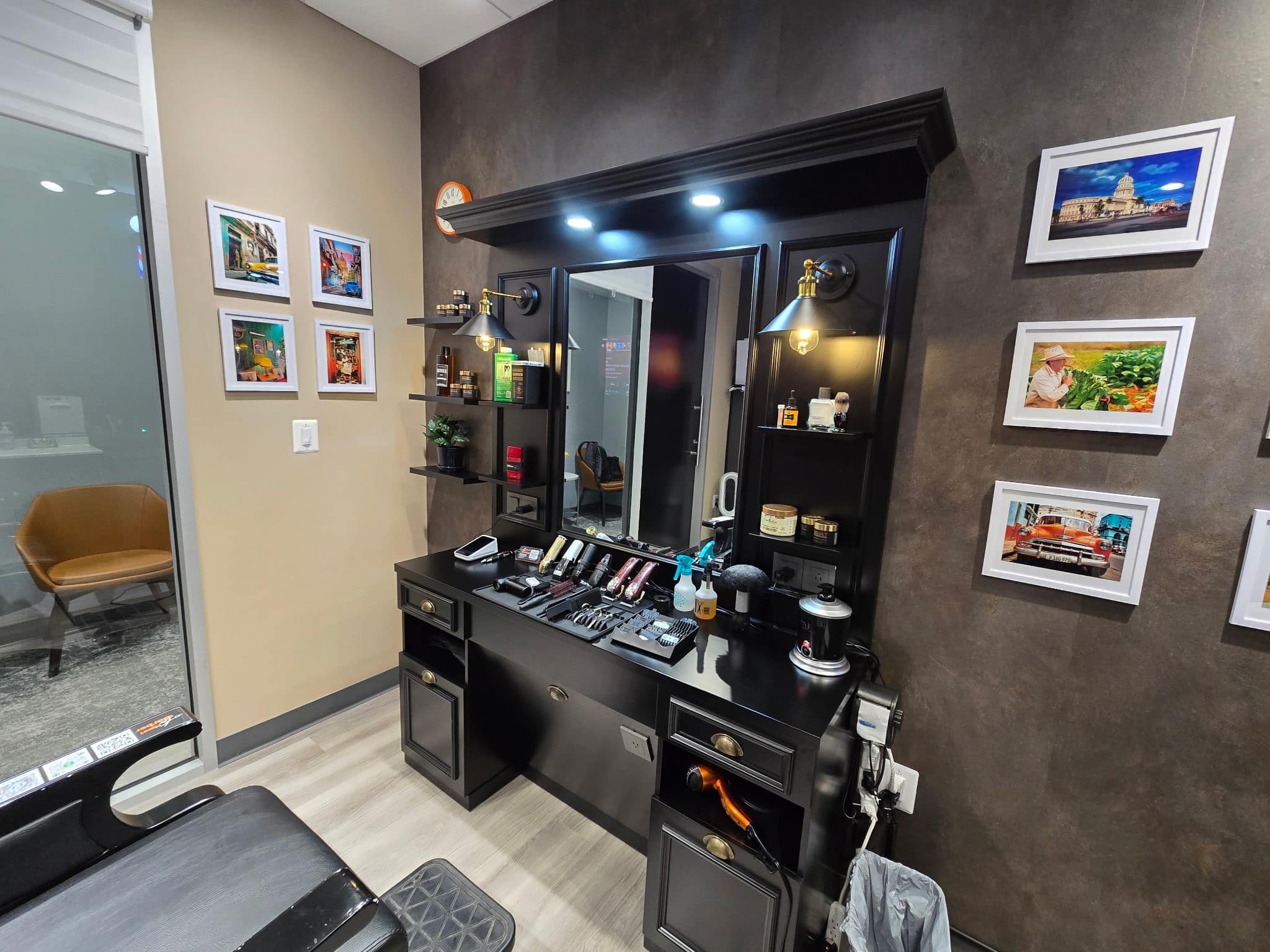 Henri's station: a black mirror unit with lit shelves of product, clippers and shears laid out on the counter, and framed prints of Cuba on the wall beside it.
